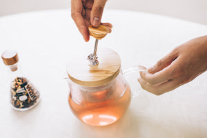 Hands lifting olive wood lid from glass teapot filled with reddish-amber tea