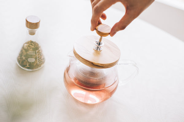 Hand placing wood and white ceramic lid on glass teapot with brewed tea