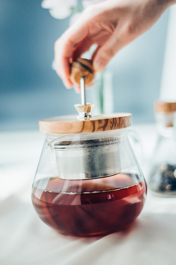 Hand removing olive wood lid from glass pot of dark steeped tea