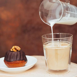 Hand pouring cream into a glass of pale beverage next to pastry.