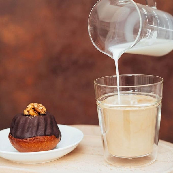 Hand pouring cream into a glass of pale beverage next to pastry.