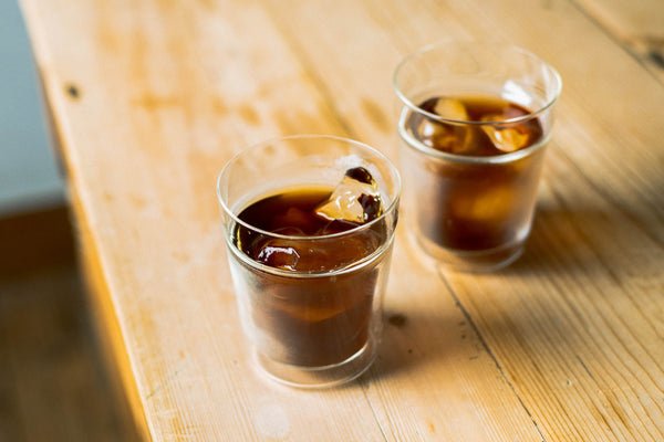 Two glasses of iced cola with ice cubes on a wooden surface in sunlight.
