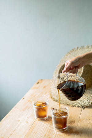 Hand pouring cola from a carafe into two iced glasses on wooden table.