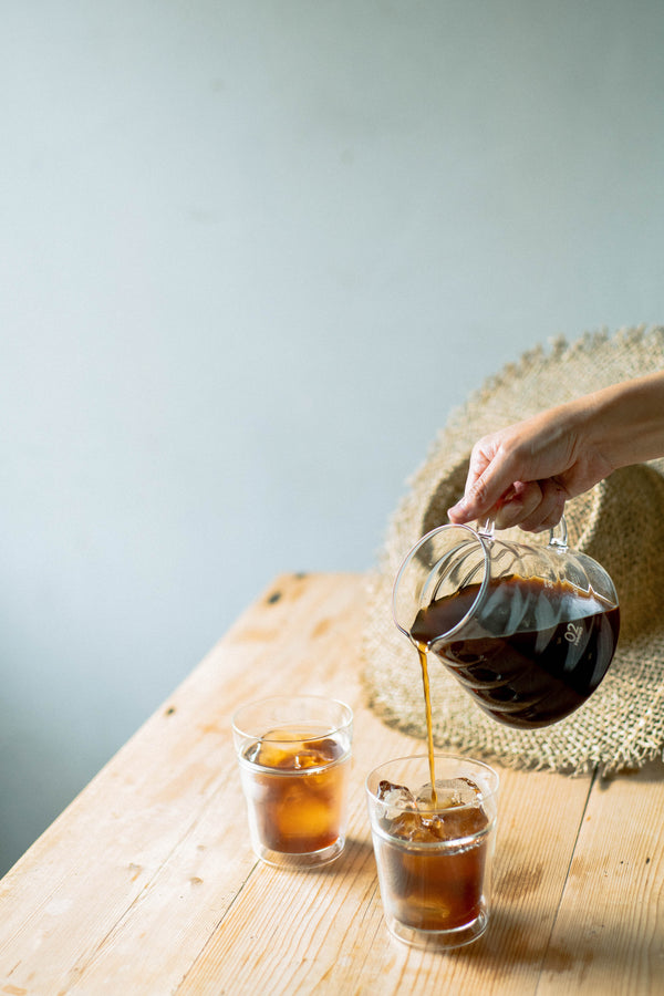 Hand pouring cola from a carafe into two iced glasses on wooden table.