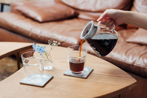 Hand pouring dark cola from a glass carafe into a cup on wooden table.