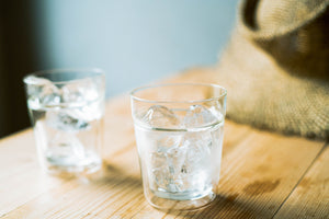 Two glasses filled with ice water on wooden surface next to fabric.