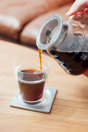 Hand pouring cola from a labeled glass container into an iced cup.
