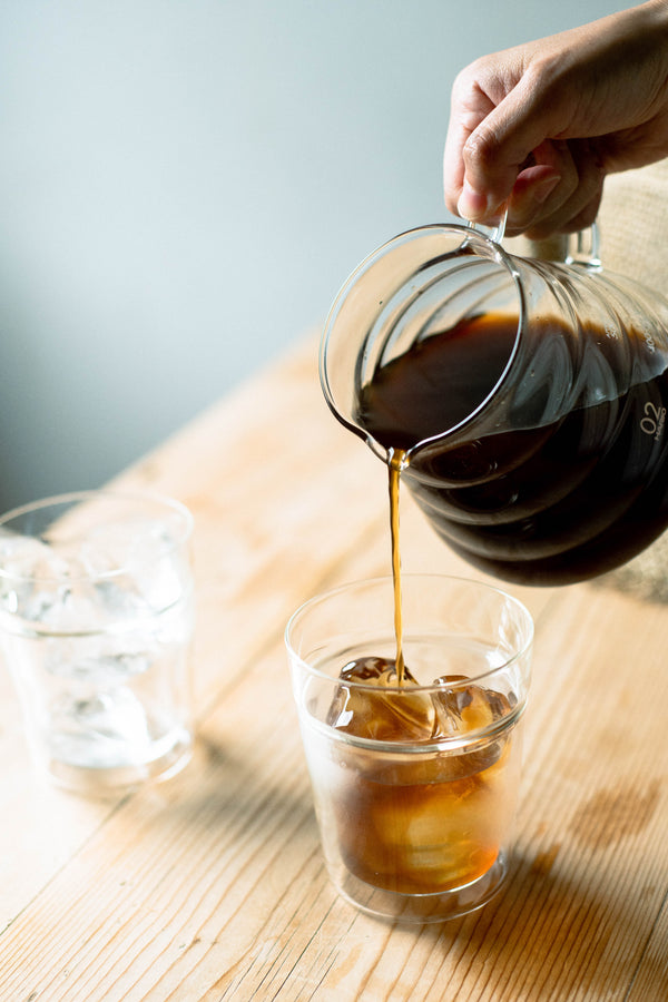 Hand pouring cola from a carafe into an iced glass on wooden table.