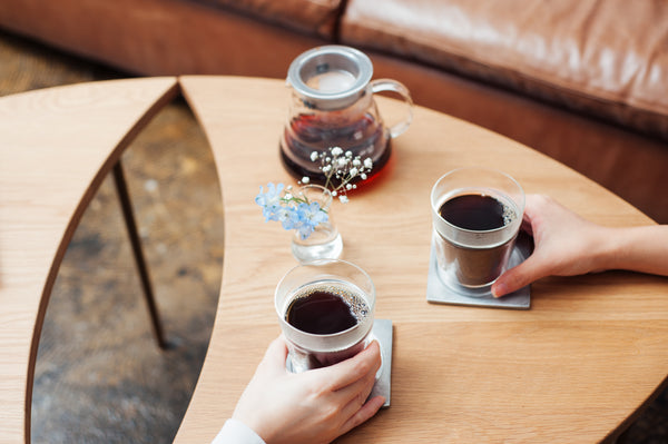 Two hands holding cups of dark cola with flowers and carafe on table.