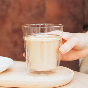 Hand holding a glass of pale liquid on a round wooden coaster.