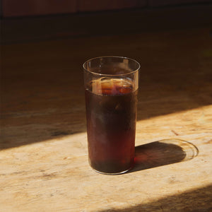 Tall glass of dark cola with ice sitting on wooden surface in sunlight.