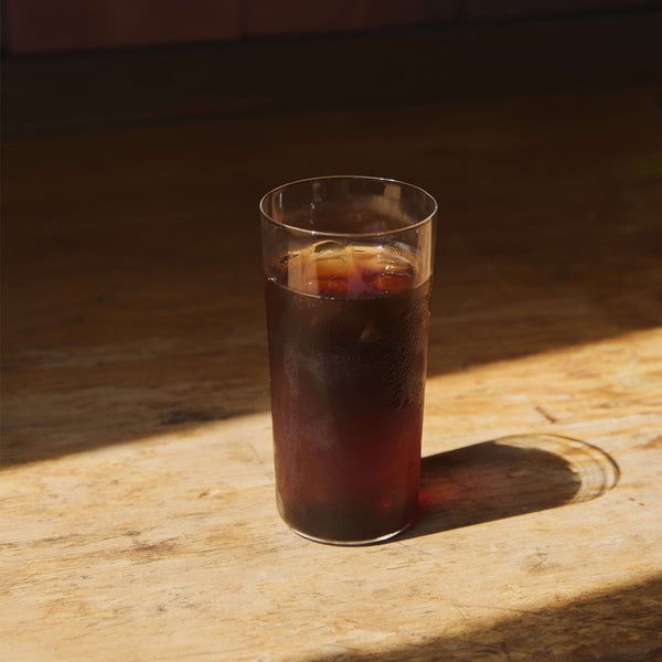 Tall glass of dark cola with ice sitting on wooden surface in sunlight.
