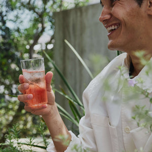 Smiling person in white shirt holding glass of pink sparkling drink outdoors.