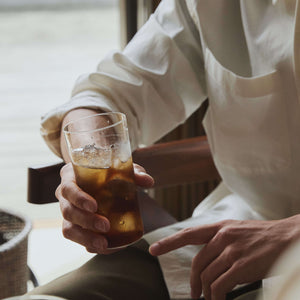 Person in cream linen shirt holding glass of cola with ice cubes.