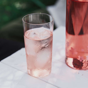 Glass of pale pink sparkling drink with ice next to pitcher on white surface.