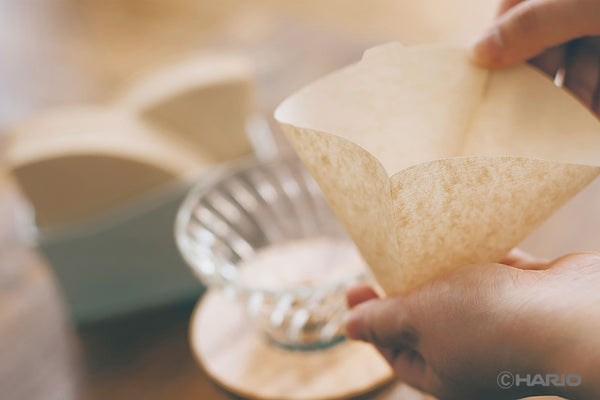 Hands holding folded cone-shaped V60 paper filter above glass dripper.