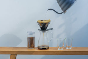 Black kettle pouring water into Hario V60 dripper over glass carafe with ground coffee and empty cups
