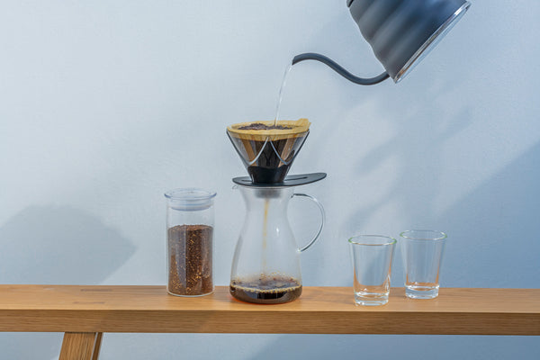 Black kettle pouring water into Hario V60 dripper over glass carafe with ground coffee and empty cups