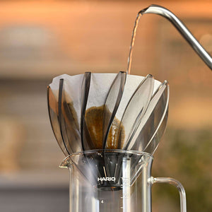V60 Suiren coffee dripper with tea bag actively brewing in glass carafe with hot water pouring from metal gooseneck kettle