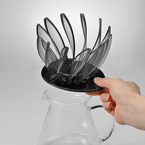 Hand holding V60 Suiren dripper over glass carafe showing transparent black petals and interior rib detail from side angle