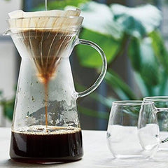 Glass pour-over coffee maker with cone filter dripping dark coffee into pitcher filled with ice cubes, outdoor setting.