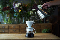 Hand pouring hot water from metal kettle into white ceramic dripper cone over glass carafe with flowers in background
