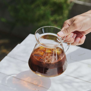 Hand holding glass coffee server with dark brewed coffee and ice cubes outdoors on white cloth