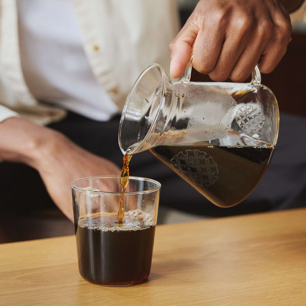 Pouring brewed coffee from glass server into cup with decorative pattern visible on both vessels