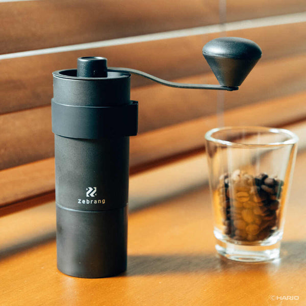 Black Zebrang coffee grinder with crank handle next to glass of brewed coffee on wooden table