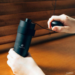 Hands holding black Zebrang coffee grinder while turning the crank handle to grind beans