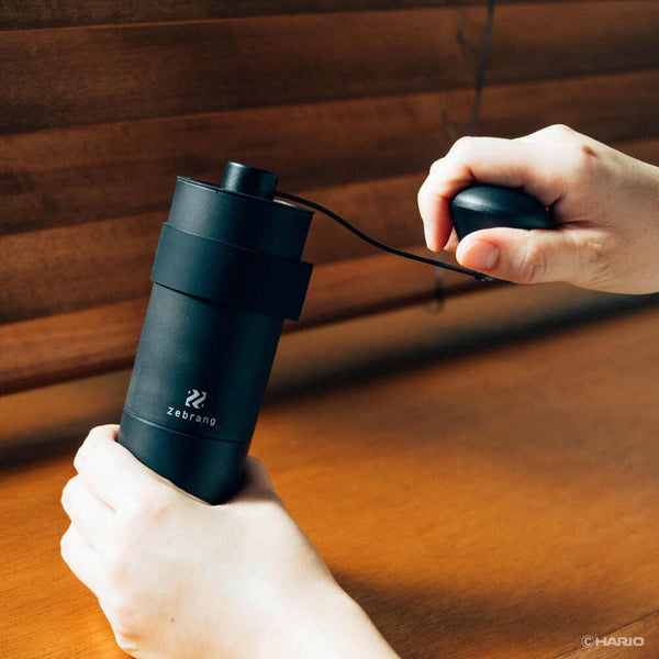 Hands holding black Zebrang coffee grinder while turning the crank handle to grind beans