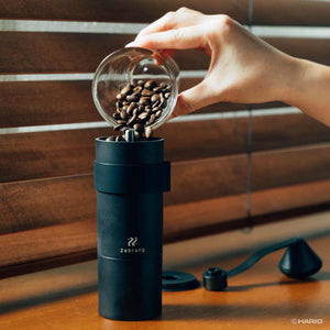 Hand pouring coffee beans from glass container into open top of black Zebrang grinder
