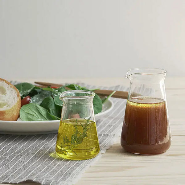 Clear glass conical pitcher filled with golden olive oil next to darker balsamic vinegar, displayed with fresh salad