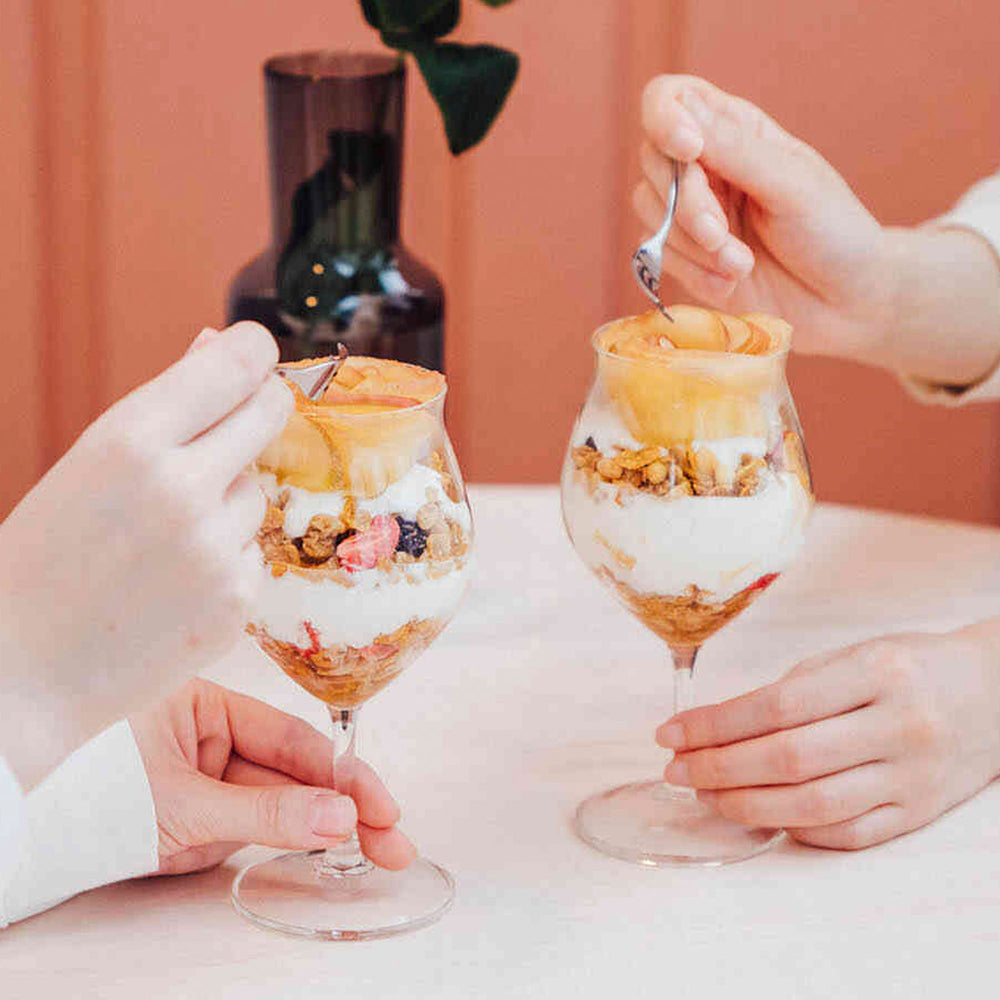 Two hands holding tulip glasses layered with granola, yogurt, and caramel drizzle against coral backdrop.