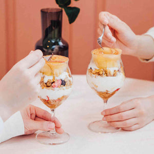 Two hands holding tulip glasses layered with granola, yogurt, and caramel drizzle against coral backdrop.