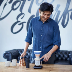 Man in blue shirt pouring water into white ceramic HARIO SWITCH dripper with gradient-striped filter cone on wooden counter